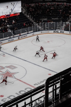 A hockey game taking place in an indoor arena with players in red and white uniforms competing on an ice rink. The arena has spectators in the stands, and a large digital screen displays the game. The rink is marked with various lines and logos.