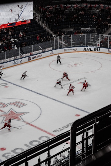 A hockey game taking place in an indoor arena with players in red and white uniforms competing on an ice rink. The arena has spectators in the stands, and a large digital screen displays the game. The rink is marked with various lines and logos.