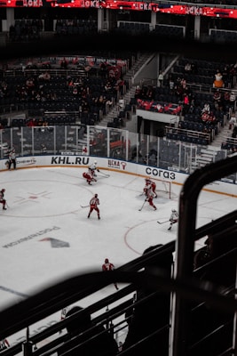 A hockey game is taking place in an indoor arena with players dressed in red and white uniforms on the ice. The stands have a sparse crowd watching the game, and banners with text and year numbers are displayed above the audience. The environment is dimly lit, giving the impression of a professional sports event.