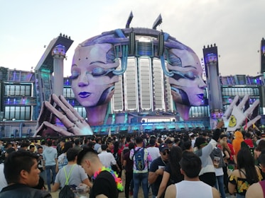 A large outdoor stage has futuristic and elaborate decorations featuring two giant sculpted faces with closed eyes, set against a backdrop of structures resembling a mix of classical and sci-fi architecture. A massive crowd of people gathers in front, many of whom appear to be enjoying a music festival.