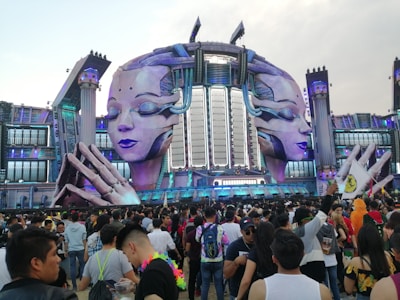 A large outdoor stage has futuristic and elaborate decorations featuring two giant sculpted faces with closed eyes, set against a backdrop of structures resembling a mix of classical and sci-fi architecture. A massive crowd of people gathers in front, many of whom appear to be enjoying a music festival.