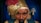 A mannequin or figure of a fortune teller, wearing a gold turban adorned with a red feather and a decorative jewel. The figure has a stern expression with thick eyebrows and a mustache. In the background, blue curtains and two lamps on the wall are visible.