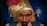 A mannequin or figure of a fortune teller, wearing a gold turban adorned with a red feather and a decorative jewel. The figure has a stern expression with thick eyebrows and a mustache. In the background, blue curtains and two lamps on the wall are visible.