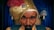 A mannequin or figure of a fortune teller, wearing a gold turban adorned with a red feather and a decorative jewel. The figure has a stern expression with thick eyebrows and a mustache. In the background, blue curtains and two lamps on the wall are visible.