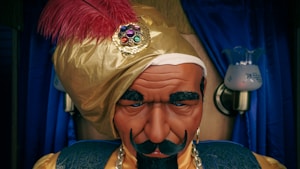A mannequin or figure of a fortune teller, wearing a gold turban adorned with a red feather and a decorative jewel. The figure has a stern expression with thick eyebrows and a mustache. In the background, blue curtains and two lamps on the wall are visible.