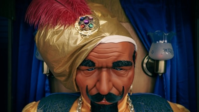 A mannequin or figure of a fortune teller, wearing a gold turban adorned with a red feather and a decorative jewel. The figure has a stern expression with thick eyebrows and a mustache. In the background, blue curtains and two lamps on the wall are visible.