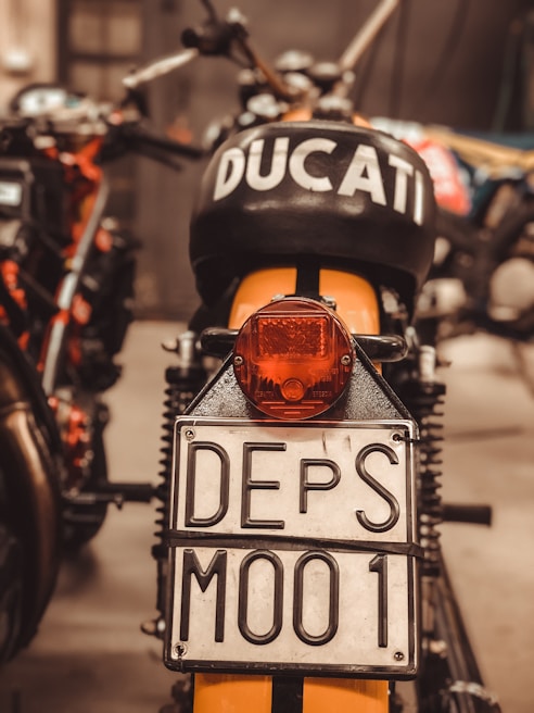 A close-up view of the rear of a motorcycle, prominently displaying a license plate marked 'DEPS M001'. The motorcycle has the word 'DUCATI' marked clearly on the black seat or tank area. The surroundings are slightly blurred but suggest a garage or workshop environment.