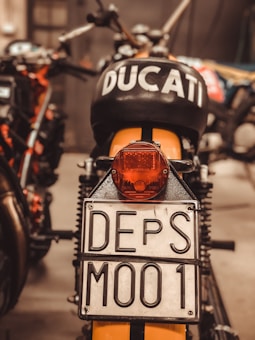 A close-up view of the rear of a motorcycle, prominently displaying a license plate marked 'DEPS M001'. The motorcycle has the word 'DUCATI' marked clearly on the black seat or tank area. The surroundings are slightly blurred but suggest a garage or workshop environment.