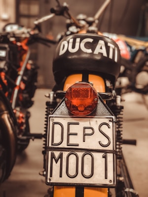 A close-up view of the rear of a motorcycle, prominently displaying a license plate marked 'DEPS M001'. The motorcycle has the word 'DUCATI' marked clearly on the black seat or tank area. The surroundings are slightly blurred but suggest a garage or workshop environment.