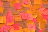 A wall covered in a vibrant assortment of sticky notes in various colors such as pink, yellow, and orange. Each note contains different handwritten messages and drawings, conveying feelings of affection and positivity. The notes feature hearts, smiley faces, and words like 'love,' 'proud,' and 'smile.'