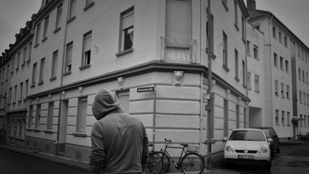 man in black jacket and black cap standing beside bicycle in grayscale photography