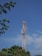 A tall communication tower stands against a clear blue sky, surrounded by a few leafy green trees. The structure is equipped with various antennas and satellite dishes, indicating its purpose for transmitting signals.