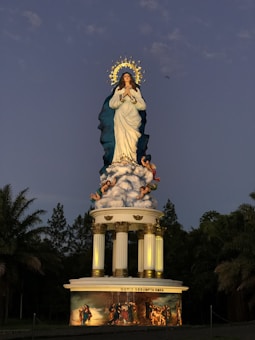 A large statue depicting a woman with a halo, wearing a white robe with a blue mantle, standing on clouds surrounded by small cherubic figures. The statue is elevated on a pedestal with ornate columns, and there is a painting on the base featuring multiple figures in a historical or religious scene. The background includes trees and a darkening sky.