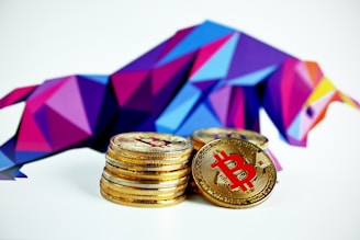 A stack of Bitcoin coins is positioned in the foreground with a vibrant, polygonal bull sculpture in the background. The bull's design features a mix of blue, purple, and pink geometric shapes.