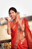 A woman wearing a vibrant red saree, accessorized with gold bangles and earrings, is smiling with an expression of joy and enthusiasm. She appears to be standing on a terrace with a blurred background of a building or structure.