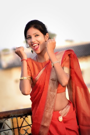 A woman wearing a vibrant red saree, accessorized with gold bangles and earrings, is smiling with an expression of joy and enthusiasm. She appears to be standing on a terrace with a blurred background of a building or structure.