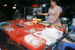 A woman is preparing food at a street food stall. There are various types of seafood in baskets, including clams and crabs. The setup is outdoors with several red and white baskets containing seafood items. A scale and some bowls are visible near the woman as she cooks.