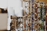 Numerous beaded necklaces in various colors, such as blue, gold, and green, hanging from a horizontal rack. The focus is on the ornate beads and intricate designs of the necklaces, with blurred bokeh lights in the background creating a warm and inviting atmosphere.