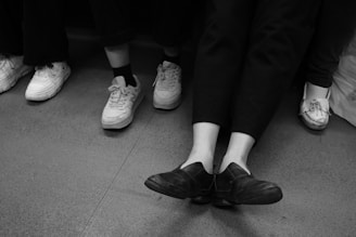 Four pairs of legs wearing different types of shoes are visible. The shoes range from casual sneakers to loafers, and the setting appears to be indoors, possibly in a subway or bus. The image is in black and white, emphasizing the textures and contrast of the floor and footwear.