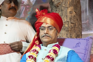 A man wearing glasses and a traditional red turban, adorned with a floral garland, sits against a tree. Beside him, another man dressed in a white traditional outfit and a smaller turban stands with both hands resting in front of him. The setting appears to be outdoors, with a tree trunk visible in the background and decorative purple cushions for support.