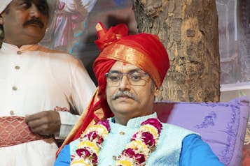 A man wearing glasses and a traditional red turban, adorned with a floral garland, sits against a tree. Beside him, another man dressed in a white traditional outfit and a smaller turban stands with both hands resting in front of him. The setting appears to be outdoors, with a tree trunk visible in the background and decorative purple cushions for support.