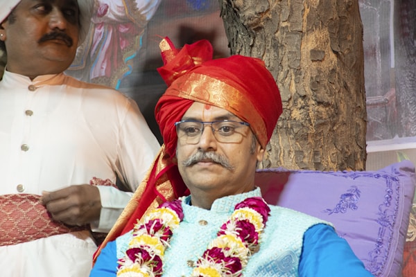 A man wearing glasses and a traditional red turban, adorned with a floral garland, sits against a tree. Beside him, another man dressed in a white traditional outfit and a smaller turban stands with both hands resting in front of him. The setting appears to be outdoors, with a tree trunk visible in the background and decorative purple cushions for support.