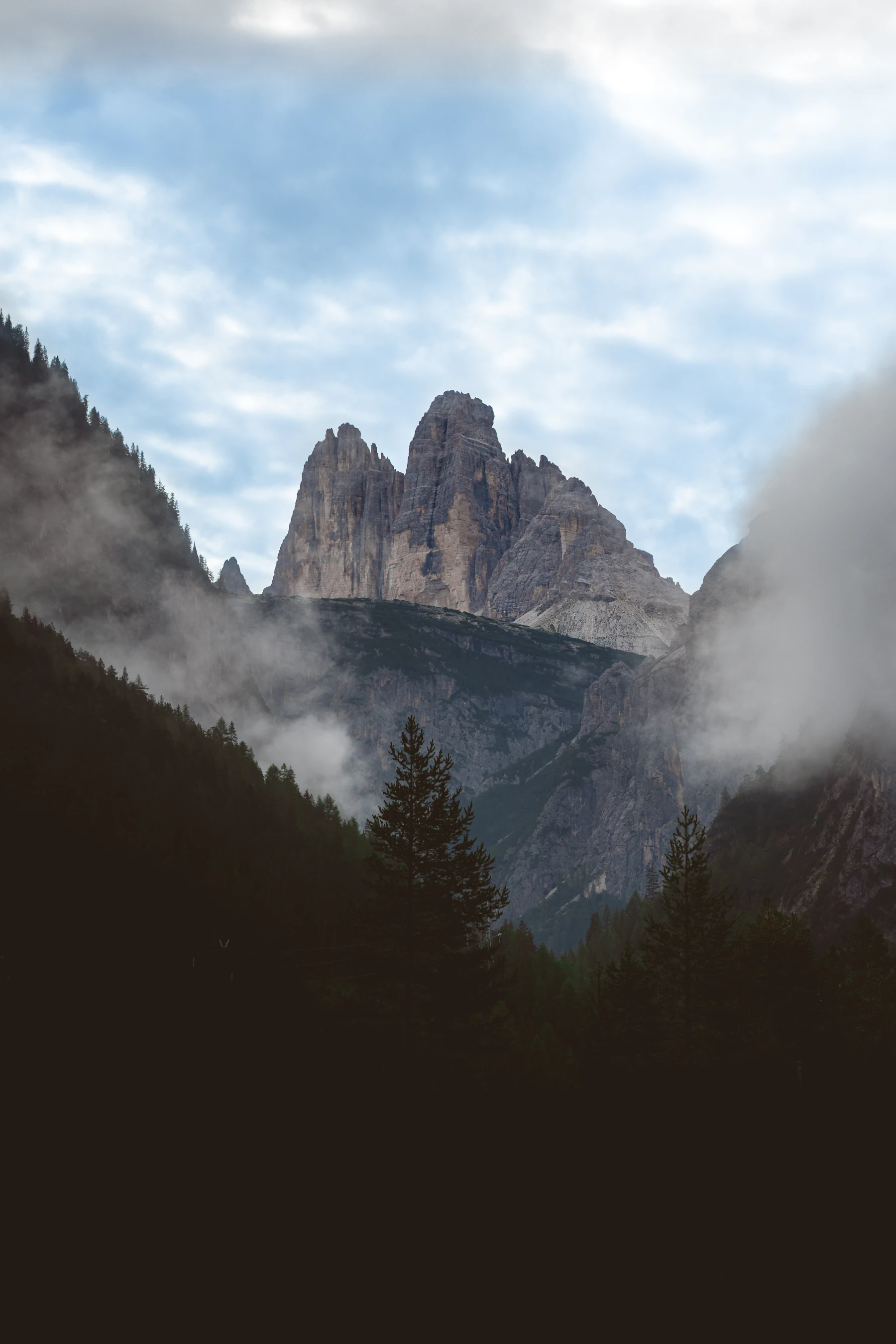Photograph of The three pale towers of Tre Cime di Lavaredo rise above dark conifers and low cloud.
