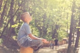 A child sitting cross-legged on a wooden stump in a serene forest setting, bathed in soft sunlight. The child appears to be in a meditative pose, with eyes closed and a peaceful expression. Sunlight filters through the trees, creating a dappled effect on the forest floor. In the background, a dirt path winds through the forest, with silhouettes of people walking.