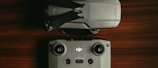 A drone is positioned next to its remote control on a wooden surface. The drone is folded, showing its propellers tucked in. The remote control has joysticks and several buttons, and both items are characterized by their grey color and modern design.