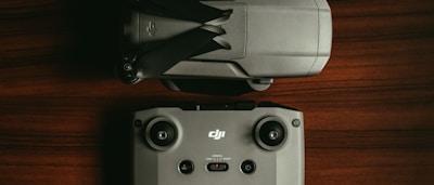 A drone is positioned next to its remote control on a wooden surface. The drone is folded, showing its propellers tucked in. The remote control has joysticks and several buttons, and both items are characterized by their grey color and modern design.