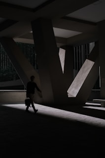 A shadowy architectural space with large, angular concrete beams casting dramatic shadows. A person is walking with a briefcase, partially illuminated by a beam of light.