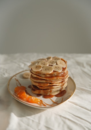 A stack of pancakes topped with sliced bananas and drizzled with syrup sits on a plate. A few slices of fresh orange are placed beside the pancakes, all set against a soft, neutral background.