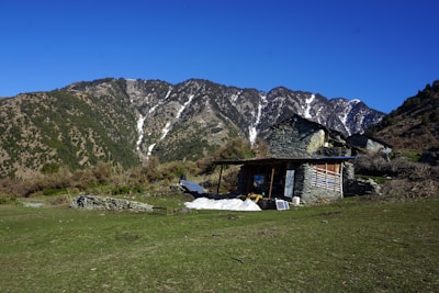 A rustic stone house with a slanted roof is situated on a grassy plain, surrounded by lush greenery and hills. Behind the house, majestic mountains with patches of snow rise against a clear blue sky. A solar panel is seen near the house, and there is a small pile of logs beside it.
