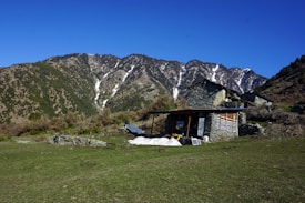 A rustic stone house with a slanted roof is situated on a grassy plain, surrounded by lush greenery and hills. Behind the house, majestic mountains with patches of snow rise against a clear blue sky. A solar panel is seen near the house, and there is a small pile of logs beside it.