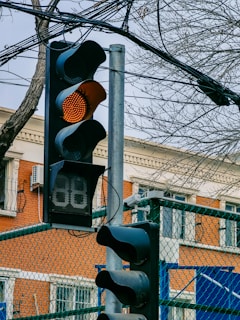 A traffic light is displaying an amber light, indicating caution for vehicles. It is mounted on a metal pole against a backdrop of urban elements including a brick building, wires, and a leafless tree. There is also a digital countdown timer below the light.