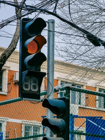A traffic light is displaying an amber light, indicating caution for vehicles. It is mounted on a metal pole against a backdrop of urban elements including a brick building, wires, and a leafless tree. There is also a digital countdown timer below the light.