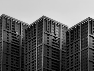 A cluster of modern high-rise buildings with a uniform architectural design, featuring numerous windows and geometric lines against an overcast sky.