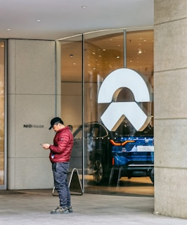 A person dressed in a red jacket and dark pants is standing outside a glass storefront, engrossed in their phone. The storefront features a large, white logo and behind the glass, a blue electric vehicle is prominently displayed in a well-lit interior with the label EC6 visible.