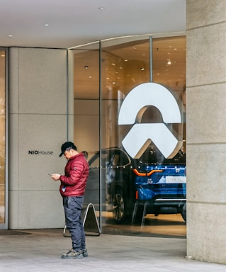 A person dressed in a red jacket and dark pants is standing outside a glass storefront, engrossed in their phone. The storefront features a large, white logo and behind the glass, a blue electric vehicle is prominently displayed in a well-lit interior with the label EC6 visible.
