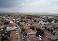 Aerial view of a residential area with numerous houses featuring red-tiled roofs. The houses are arranged in a grid-like pattern with several trees interspersed between them. A curved road runs through the area, with a few vehicles visible on it. The background features open fields and a mountain range with a cloudy sky overhead.