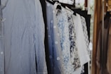 A collection of shirts hanging on a clothing rack. The shirts feature various patterns, including stripes and floral designs, and are mostly in shades of blue and white. The scene is well-lit with natural light coming from a nearby window.