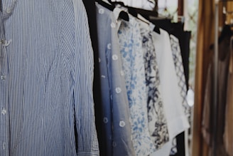 A collection of shirts hanging on a clothing rack. The shirts feature various patterns, including stripes and floral designs, and are mostly in shades of blue and white. The scene is well-lit with natural light coming from a nearby window.