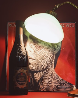 An intricately designed wine bottle stands in front of a red background featuring stylized, abstract artwork of a face. A vintage-style lamp illuminates the scene, casting a warm glow over the arrangement.