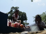 Two people are outdoors, potentially involved in a cleanup or meal distribution activity. They are surrounded by cups and bags on a surface. One person is wearing a red bandana and gloves. Trees and a clear sky form the background.