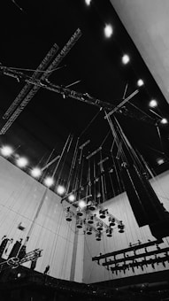 A large industrial space is filled with a complex network of overhead lighting rigs and suspended equipment. The scene features numerous spotlights hanging from the ceiling, supported by a system of wires and metal trusses, creating a sense of height and scale. The overall ambiance is utilitarian, with visible structural elements and functional design.