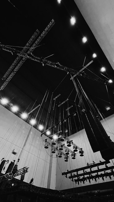 A large industrial space is filled with a complex network of overhead lighting rigs and suspended equipment. The scene features numerous spotlights hanging from the ceiling, supported by a system of wires and metal trusses, creating a sense of height and scale. The overall ambiance is utilitarian, with visible structural elements and functional design.