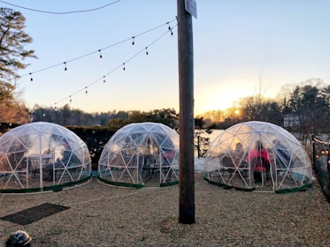 A scenic outdoor setting with three transparent geodesic domes situated on a gravel surface near a body of water. Each dome contains tables and chairs, hinting at a dining setup. String lights hang above, adding ambiance as the sun sets over a tree-lined horizon.