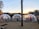 A scenic outdoor setting with three transparent geodesic domes situated on a gravel surface near a body of water. Each dome contains tables and chairs, hinting at a dining setup. String lights hang above, adding ambiance as the sun sets over a tree-lined horizon.