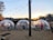 A scenic outdoor setting with three transparent geodesic domes situated on a gravel surface near a body of water. Each dome contains tables and chairs, hinting at a dining setup. String lights hang above, adding ambiance as the sun sets over a tree-lined horizon.