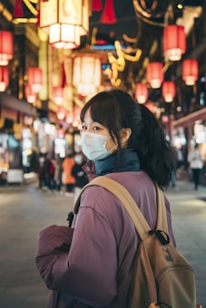 A young woman with a face mask is walking through a vibrant street adorned with hanging red lanterns. She is wearing a purple coat and carrying a beige backpack. The street is bustling with people in the background, and the lighting creates a warm, festive atmosphere.